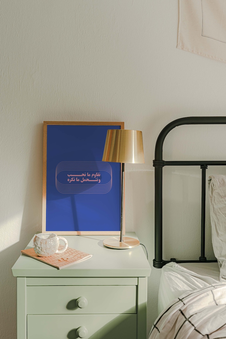 Pu&ograve; includere: Una stampa incorniciata con sfondo blu e testo arabo rosa che recita "Naqoum ma na'hib wa nas'hal ma na'krez". La stampa &egrave; su un comodino accanto a una lampada dorata e una tazza di ceramica bianca.
