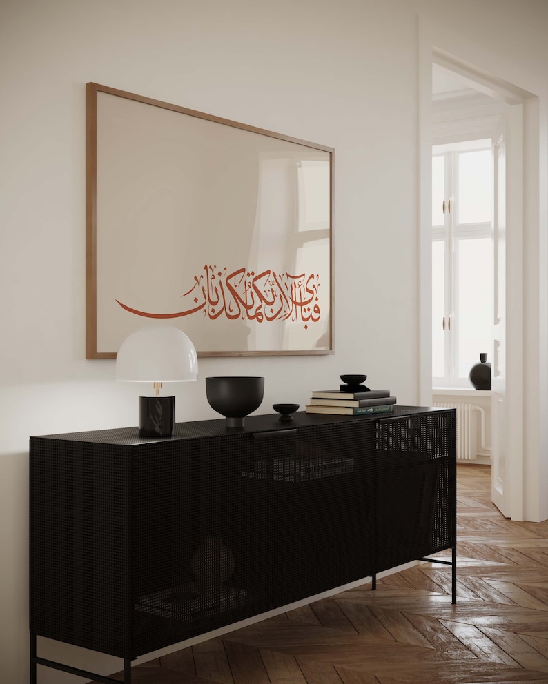May include: A black metal cabinet with a mesh front and two doors. The cabinet is sitting on a wooden floor. A framed print with Arabic calligraphy hangs on the wall above the cabinet. A lamp and decorative bowls are on top of the cabinet.