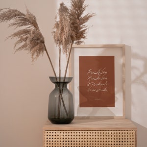 May include: A wooden frame with a brown background and white calligraphy text in Arabic. The frame is sitting on a light wood table with a woven rattan drawer. A vase with dried pampas grass is on the table.