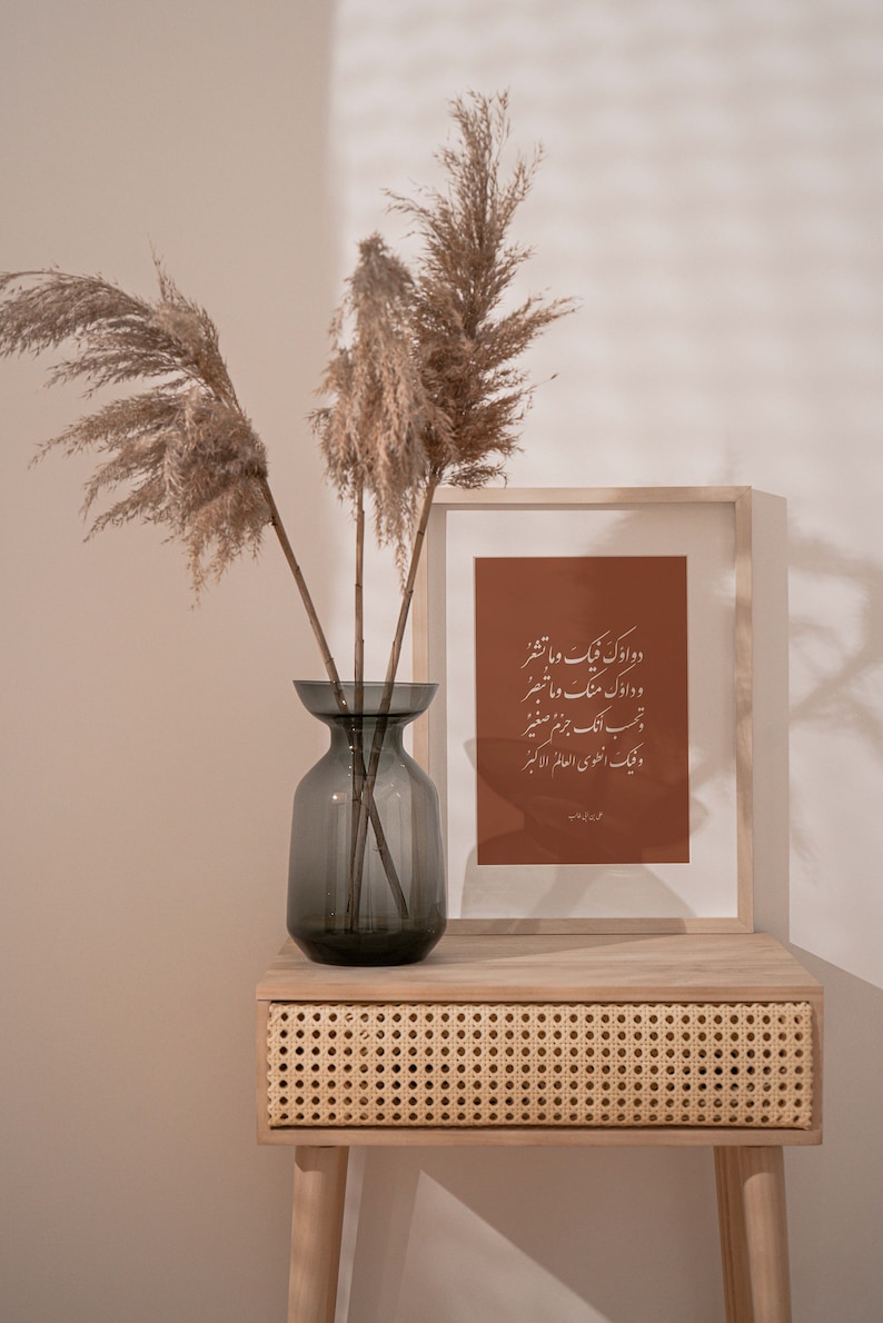 May include: A wooden frame with a brown background and white calligraphy text in Arabic. The frame is sitting on a light wood table with a woven rattan drawer. A vase with dried pampas grass is on the table.