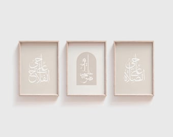 May include: Three framed art prints with beige backgrounds and light wood frames. Each print features white Arabic calligraphy. The central print has an arched design. The other two prints have rectangular designs. The prints are displayed against a white wall.