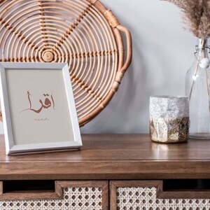 Puede incluir: Un marco de fotos blanco con una impresi&oacute;n de caligraf&iacute;a &aacute;rabe marr&oacute;n que dice "Leer" en ingl&eacute;s. El marco est&aacute; sobre una mesa de madera con una bandeja de mimbre tejido y un jarr&oacute;n de vidrio con hierba de la pampa seca detr&aacute;s.