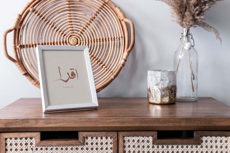 Puede incluir: Un marco de fotos blanco con una impresi&oacute;n de caligraf&iacute;a &aacute;rabe marr&oacute;n que dice "Leer" en ingl&eacute;s. El marco est&aacute; sobre una mesa de madera con una bandeja de mimbre tejido y un jarr&oacute;n de vidrio con hierba de la pampa seca detr&aacute;s.