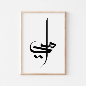 Puede incluir: Impresión artística enmarcada con elegante caligrafía árabe negra sobre fondo blanco. La obra de arte se presenta en un marco de madera clara, añadiendo un toque de calidez. El diseño minimalista es adecuado para varios estilos de interior.