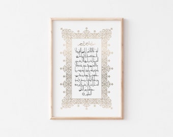 May include: Framed artwork featuring Arabic calligraphy in black ink on a white background. The text is surrounded by an ornate gold border and framed in a light wood frame. The artwork is displayed against a white wall.