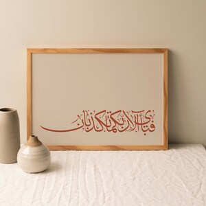 May include: A framed print with a brown wooden frame. The print features a red Arabic calligraphy design on a white background. The print is sitting on a white table with two ceramic vases.