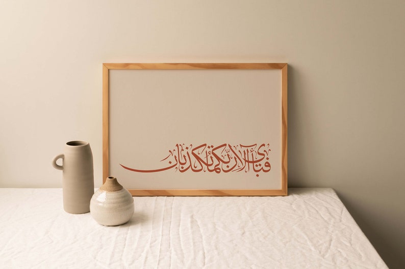 May include: A framed print with a brown wooden frame. The print features a red Arabic calligraphy design on a white background. The print is sitting on a white table with two ceramic vases.
