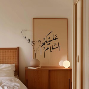 May include: A beige bedroom with a wooden headboard, a bedside table with a lamp, and a framed print with Arabic calligraphy that reads "عليكَ السلام" on the wall.
