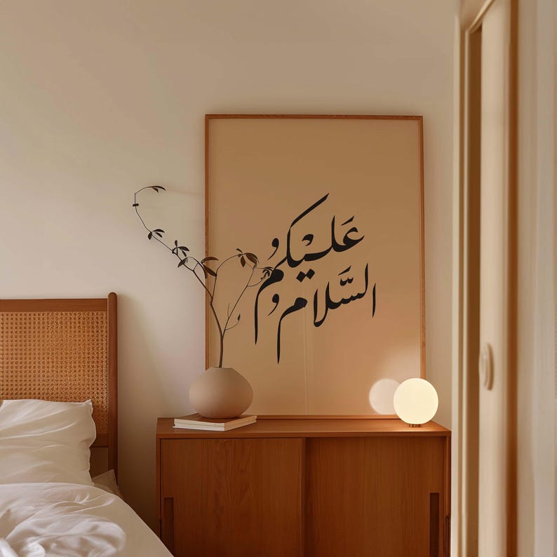 May include: A beige bedroom with a wooden headboard, a bedside table with a lamp, and a framed print with Arabic calligraphy that reads "عليكَ السلام" on the wall.