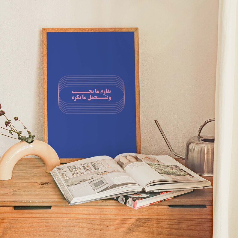 Pu&ograve; includere: Una stampa incorniciata con sfondo blu e testo rosa che recita "نلوم ما نتعب ونحسب ما نكره". La stampa &egrave; appoggiata su un tavolo di legno con un libro e un annaffiatoio.