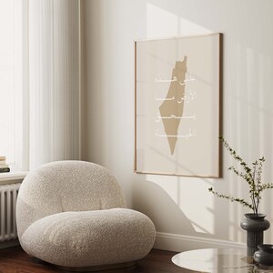 May include: A beige armchair with a fluffy texture sits in a room with a framed print of the outline of Palestine in beige with Arabic text in white. The print is hanging on a white wall. A small vase with a green plant and a black vase are on a glass table.