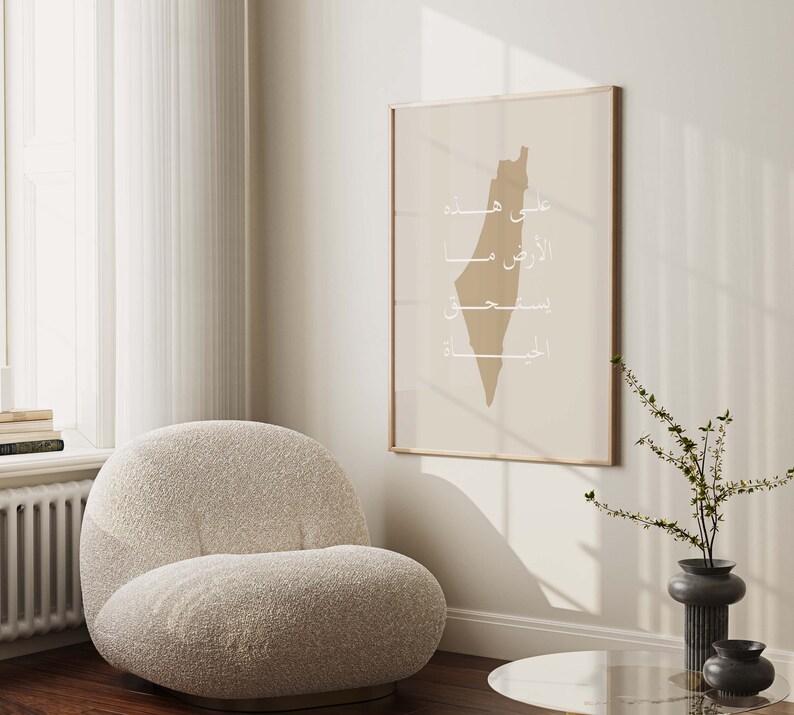 May include: A beige armchair with a fluffy texture sits in a room with a framed print of the outline of Palestine in beige with Arabic text in white. The print is hanging on a white wall. A small vase with a green plant and a black vase are on a glass table.