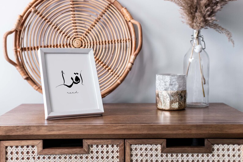 Puede incluir: Una impresi&oacute;n enmarcada en blanco con caligraf&iacute;a &aacute;rabe negra que dice "أقرأ" y la palabra "leer" en ingl&eacute;s debajo. La impresi&oacute;n est&aacute; sobre una mesa de madera con una bandeja de mimbre tejido y un jarr&oacute;n de vidrio con hierba de la pampa seca detr&aacute;s.