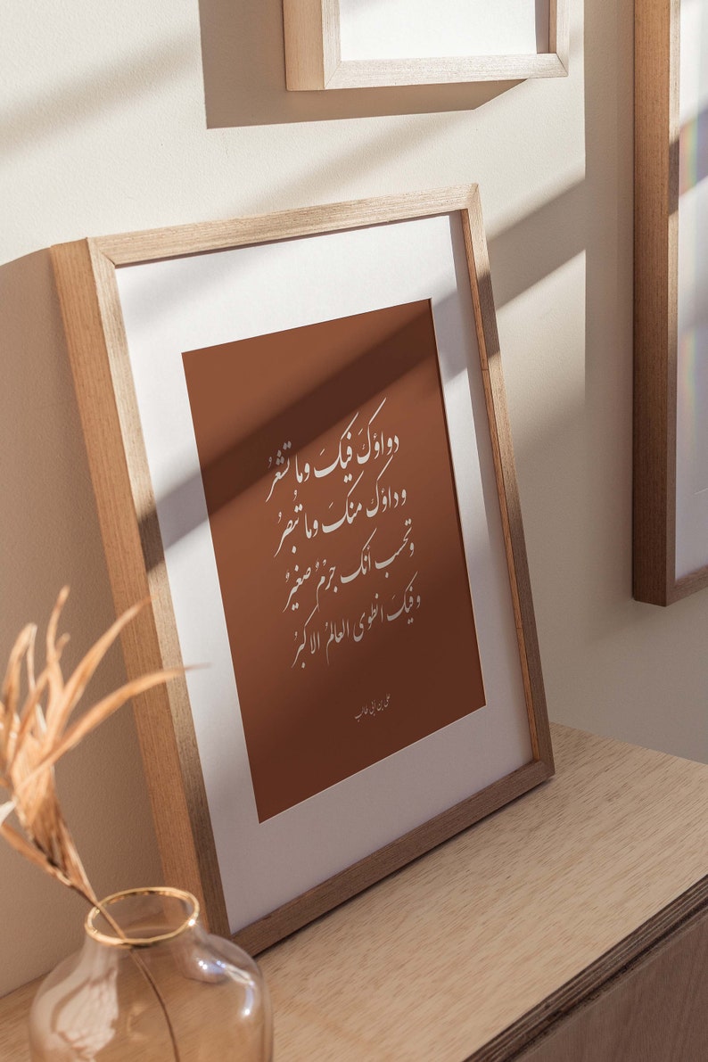 May include: A framed print with a brown background and white Arabic calligraphy. The text reads "Two good deeds are better than a thousand prayers." The print is in a light wood frame and is sitting on a wooden surface.