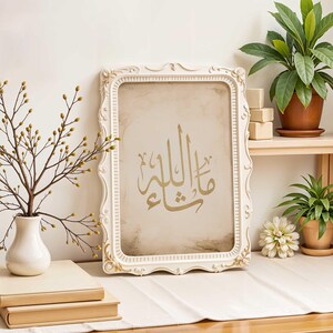 May include: A white ornate frame with a gold Arabic calligraphy print on a beige background. The frame is sitting on a wooden table with books and a vase of branches.
