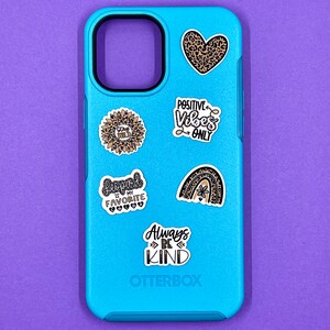 May include: A blue Otterbox phone case with various stickers. The stickers include a sunflower with the text "Stay Wild", a leopard print heart, a rainbow with a flower, a sticker that says "Leopard is my favorite", and a sticker that says "Positive Vibes Only".