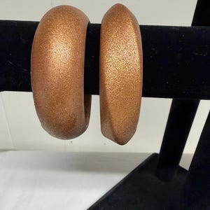 May include: Two copper-colored bangles with a glittery finish are displayed on a black jewelry stand. The bangles have a rounded, thick design, and the metallic finish catches the light, creating a shimmering effect. The jewelry is set against a white background.
