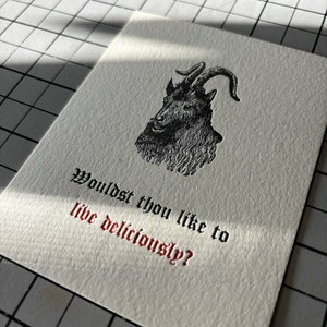 May include: A cream-colored card featuring a black illustration of a goat's head with curved horns. The text "Wouldst thou like to live deliciously?" is printed in black and red gothic font.