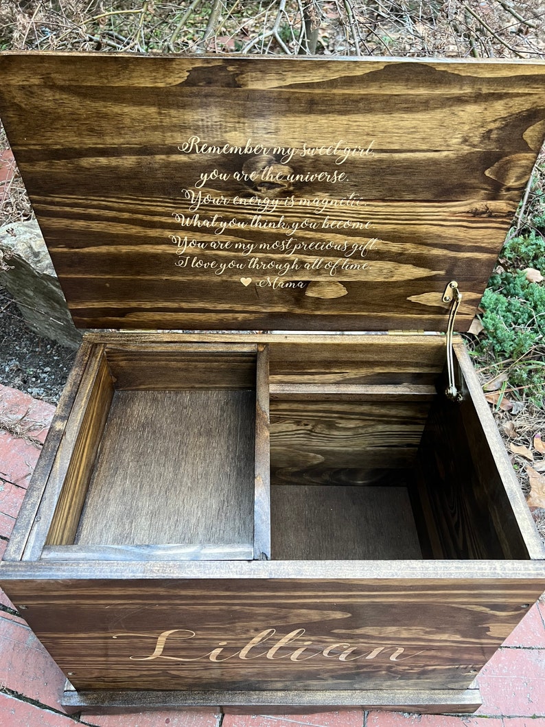 Puede incluir: Una caja de madera para recuerdos con una tapa con bisagras. La tapa est&aacute; abierta y presenta un mensaje grabado: "Remember my sweet girl, you are the universe. Your energy is magnetic. What you think you become. You are my most precious gift. I love you through all of time. Mama." La caja est&aacute; cerrada y tiene el nombre "Lillian" grabado en la parte delantera.