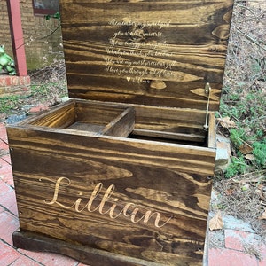 Puede incluir: Un cofre de madera para juguetes con una tapa con bisagras. La tapa est&aacute; abierta y tiene una inscripci&oacute;n dorada que dice "Recuerda mi dulce ni&ntilde;a, t&uacute; eres el universo. Tu energ&iacute;a es magn&eacute;tica. Lo que piensas, te conviertes. Eres mi regalo m&aacute;s preciado. Te amo a trav&eacute;s de todo el tiempo. Mam&aacute;." El frente del cofre est&aacute; grabado con el nombre "Lillian".