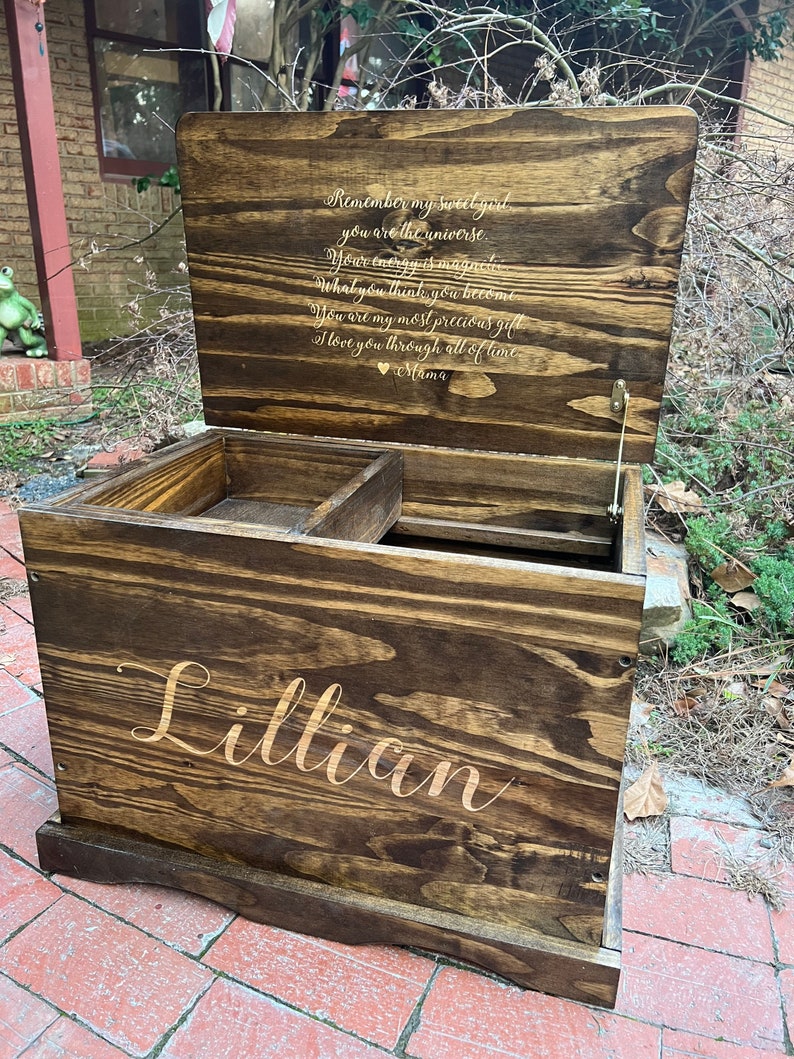 Puede incluir: Un cofre de madera para juguetes con una tapa con bisagras. La tapa est&aacute; abierta y tiene una inscripci&oacute;n dorada que dice "Recuerda mi dulce ni&ntilde;a, t&uacute; eres el universo. Tu energ&iacute;a es magn&eacute;tica. Lo que piensas, te conviertes. Eres mi regalo m&aacute;s preciado. Te amo a trav&eacute;s de todo el tiempo. Mam&aacute;." El frente del cofre est&aacute; grabado con el nombre "Lillian".