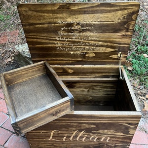 Puede incluir: Un cofre de madera para juguetes con una tapa con bisagras y un compartimento m&aacute;s peque&ntilde;o en la parte delantera. La tapa est&aacute; abierta y tiene un mensaje personalizado grabado en el interior: "Remember my sweet girl you are the universe. Your energy is magnetic. What you think, you become. You are my most precious gift. I love you through all of time. Mama." El cofre est&aacute; etiquetado con "Lillian" en la parte delantera.