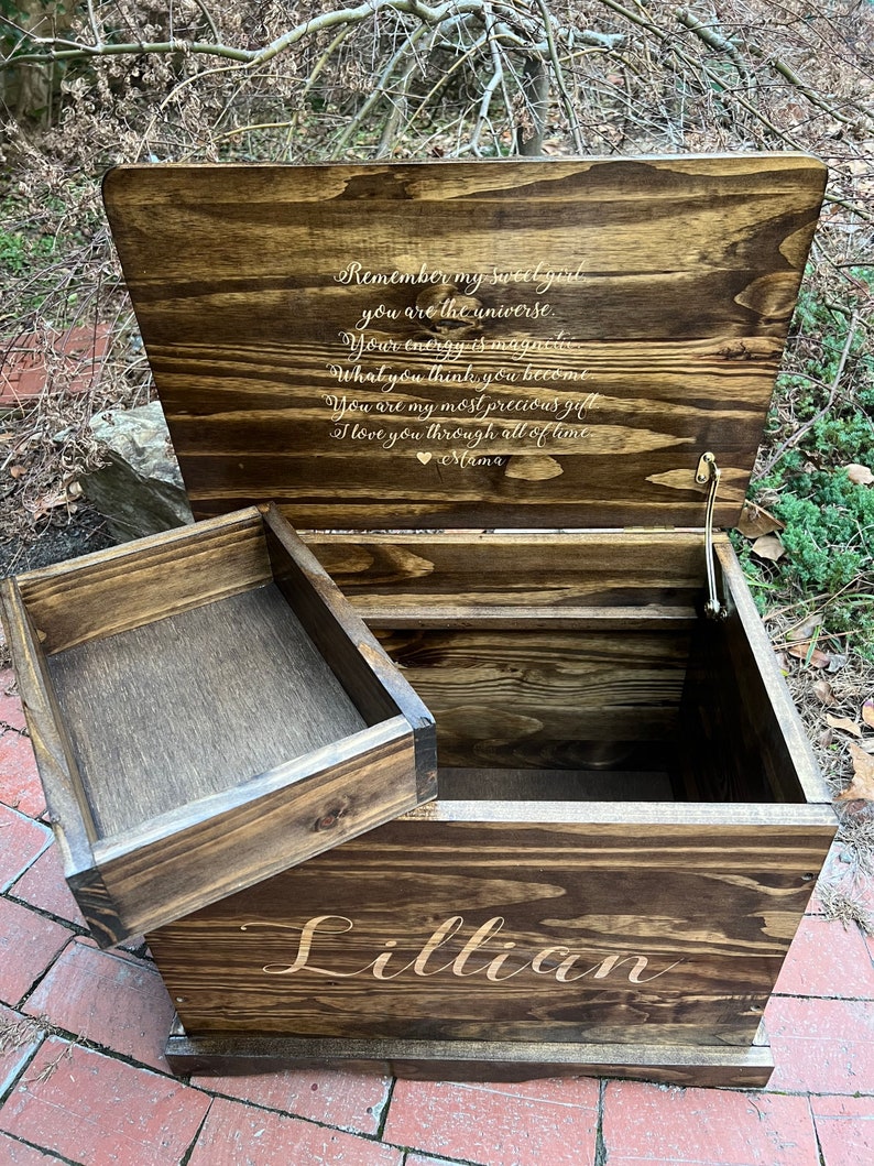 Puede incluir: Un cofre de madera para juguetes con una tapa con bisagras y un compartimento m&aacute;s peque&ntilde;o en la parte delantera. La tapa est&aacute; abierta y tiene un mensaje personalizado grabado en el interior: "Remember my sweet girl you are the universe. Your energy is magnetic. What you think, you become. You are my most precious gift. I love you through all of time. Mama." El cofre est&aacute; etiquetado con "Lillian" en la parte delantera.