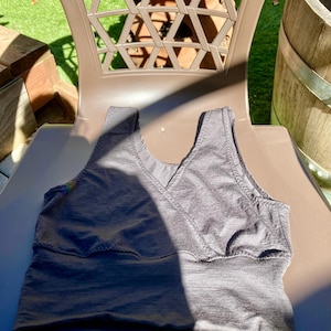 May include: A dark gray tank top with a V-neck design, resting on a brown plastic chair. The top has a textured appearance and a stitched detail along the neckline and under the bust. The lighting creates shadows and highlights on the fabric.