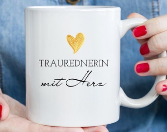 May include: White ceramic mug with a gold heart and the text "TRAUREDNERIN mit Herz" in black.