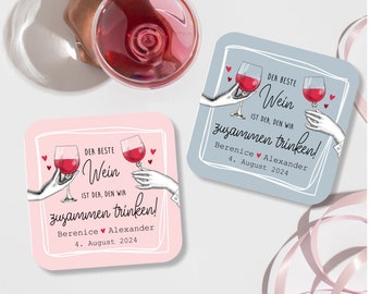 May include: Two pink and blue square coasters with a white border. Each coaster features a hand-drawn illustration of two hands holding wine glasses with the text "Der Beste Wein ist der, den wir zusammen trinken!" and the names "Berenice Alexander" and the date "4. August 2024".