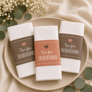 May include: Three white folded napkins with decorative bands in brown and orange, each featuring a heart and the text "Nur für Freudentränen". The napkins are arranged on a beige plate, with floral and greenery accents.