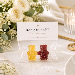 Puede incluir: Una bolsa transparente de celofán contiene dos ositos de goma, uno amarillo y otro rojo, tomados de la mano. Una tarjeta blanca encima dice "HAND IN HAND. Ein Leben lang." con un símbolo de corazón. El fondo incluye flores, una vela y anillos de boda.