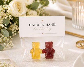 May include: A clear plastic bag containing two gummy bears, one yellow and one red, holding hands. The bag is sealed with a white paper topper featuring the text "HAND IN HAND. Ein Leben lang." with small heart icons above and below the text.
