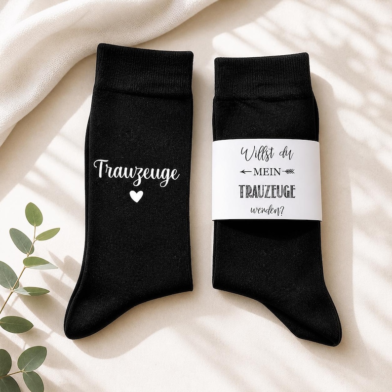 May include: A pair of black socks. One sock features the word "Trauzeuge" in white script with a small white heart. The other sock has a white band with the text "Willst du MEIN TRAUZEUGE werden?" in black.