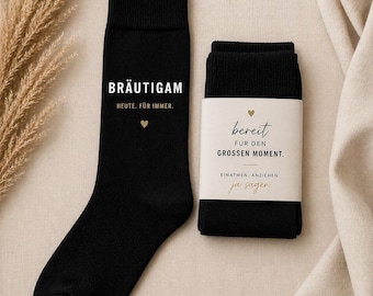 May include: Black socks with white text that reads "BRÄUTIGAM" and "HEUTE. FÜR IMMER." A gold heart is below the text. A second pair of black socks is wrapped with a label that reads "bereit FÜR DEN GROSSEN MOMENT. EINATMEN. ANZIEHEN. ja sagen."