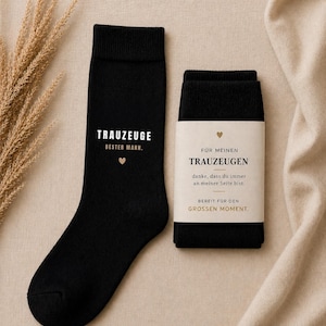 Könnte beinhalten: Schwarze Socken mit weißem Text "TRAUZEUGE BESTER MANN." und einem kleinen Herz. Ein zweites Paar schwarze Socken ist mit einem Etikett mit der Aufschrift "FÜR MEINEN TRAUZEUGEN" und anderem Text umwickelt.