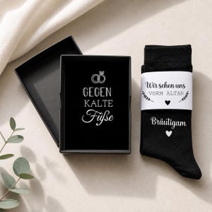 Puede incluir: Un par de calcetines negros con una banda de papel blanca que incluye el texto "Wir sehen uns VORM ALTAR" y "Bräutigam" con corazones, presentados en una caja de regalo negra con el texto "GEGEN KALTE FÜßE" y dos anillos de boda.
