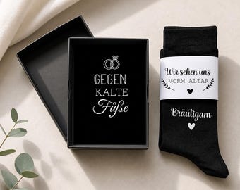 May include: A pair of black socks with a white paper band featuring the text "Wir sehen uns VORM ALTAR" and "Bräutigam" with heart icons, presented in a black gift box with the text "GEGEN KALTE FÜßE" and a graphic of two wedding rings.