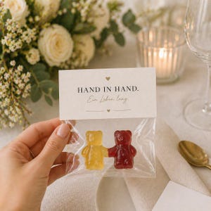 May include: A clear plastic bag containing two gummy bears, one yellow and one red, joined at the hands. The bag is sealed with a white paper topper featuring the text "HAND IN HAND." and "Ein Leben lang" with small heart icons.