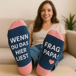 May include: A pair of navy blue and pink socks with the text "WENN DU DAS HIER LIEST" and "FRAG PAPA!" printed in white. A pink heart is also printed on one of the socks. The socks are worn by a person sitting on a sofa.