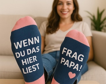 May include: A pair of navy blue and pink socks with the text "WENN DU DAS HIER LIEST" and "FRAG PAPA!" printed in white. A pink heart is also printed on one of the socks. The socks are worn by a person sitting on a couch.