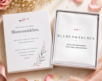 May include: A white gift box with the text "Blumenmädchen" in black, with a pink heart and arrow design. The box is open, revealing a white cloth with the same text. Floral and leaf accents in pink and gray.