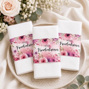 May include: Three white napkins with a floral band featuring pink and purple flowers and the text "Für deine Freudentränen". The napkins are arranged on a light-colored plate, with flowers in the background.