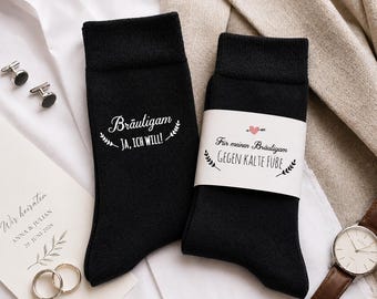 May include: Two black socks with white text, one reading "Bräutigam JA, ICH WILL!" and the other with a label saying "Für meinen Bräutigam GEGEN KALTE FUBE". The image includes a wedding invitation, rings, cufflinks, and a watch.