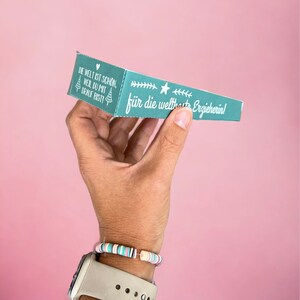 May include: A teal blue box with white text that reads "Die Welt ist schon, weil du mit drauf bist!" and "f&uuml;r die weltbeste Erzieherin!" The box is held in a hand with a colorful bracelet.