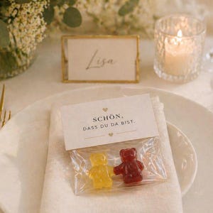 May include: A clear plastic bag containing one yellow and one red gummy bear, topped with a white paper header featuring the text "SCH&Ouml;N, DASS DU DA BIST." and two small heart icons, placed on a white napkin on a white ceramic plate.