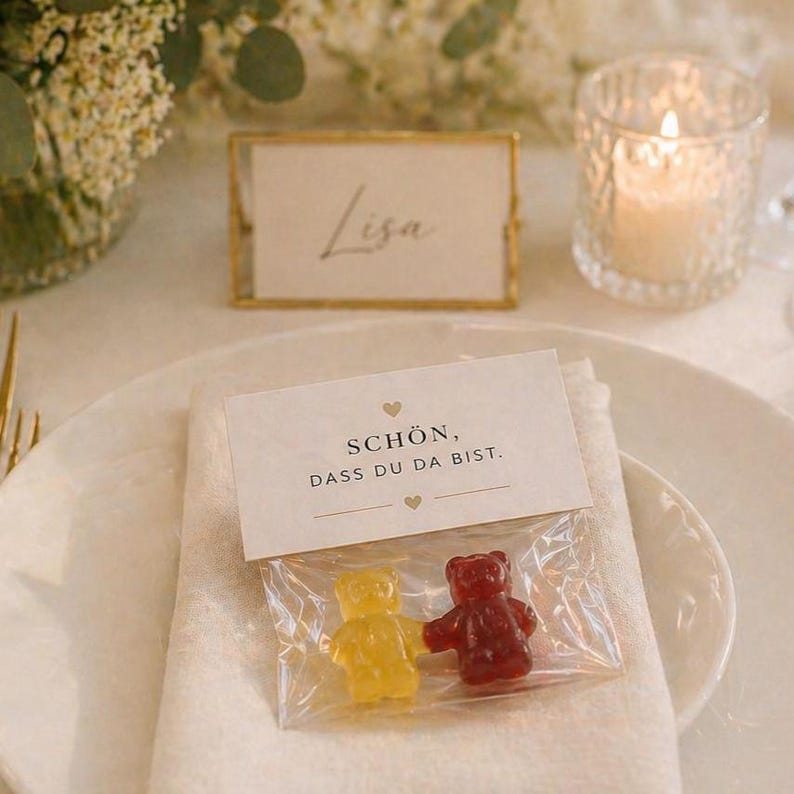 May include: A clear plastic bag containing one yellow and one red gummy bear, topped with a white paper header featuring the text "SCH&Ouml;N, DASS DU DA BIST." and two small heart icons, placed on a white napkin on a white ceramic plate.