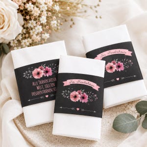 May include: Three white folded napkins with black bands featuring floral designs and text. The bands have pink flowers and text in German. One band reads "Alle Tränen dieser Welt, sollten Freudentränen set".