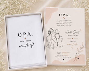 May include: A white gift box with a matching folded cloth inside, featuring the text "OPA. Für immer mein Held" in black script. The box next to it has a drawing of a couple and German text. The overall theme is a wedding gift.
