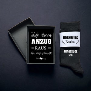 May include: A black gift box with a black interior, next to a pair of black socks with a white band. The box contains a black card with white text that reads "Hole deinen ANZUG - RAUS!" and "Du wirst gebraucht!" The socks say "HOCHZEITS Socken" and "TRAUZEUGE".